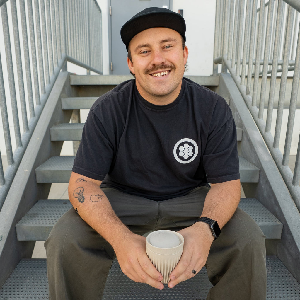 Man sitting on stairs holding a cup, wearing a black t-shirt with a logo and green pants.