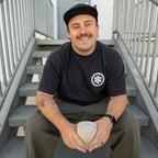 Man sitting on stairs holding a cup, wearing a black t-shirt with a logo and green pants.