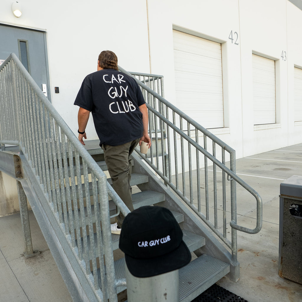 Person walking up stairs with 'Car Guy Club' shirt and cap in a building setting
