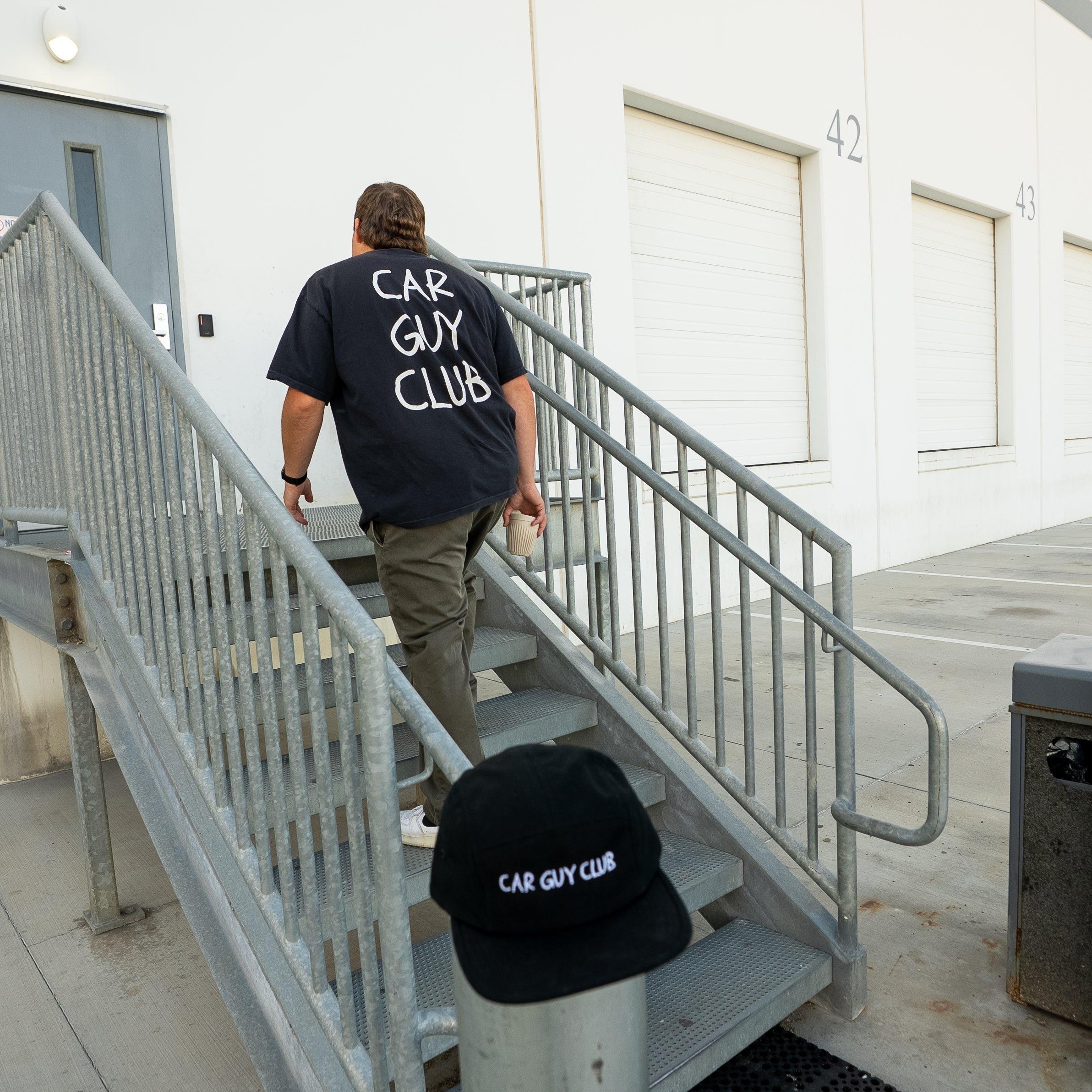 Person walking up stairs with 'Car Guy Club' shirt and cap in a building setting
