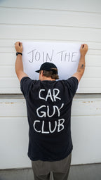 Person wearing a black t-shirt with 'Car Guy Club' text, standing in front of a white garage door with a sign that says 'Join The Car Guy Club'.