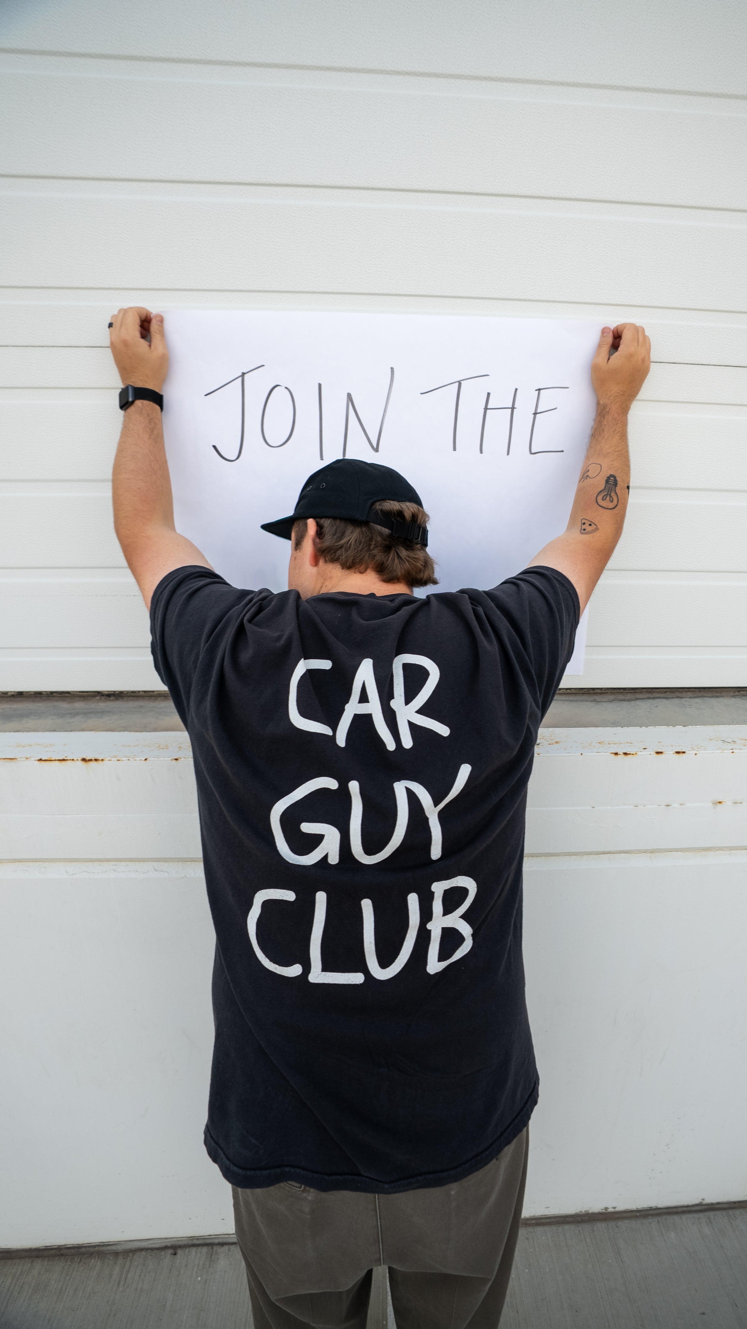 Person wearing a black t-shirt with 'Car Guy Club' text, standing in front of a white garage door with a sign that says 'Join The Car Guy Club'.