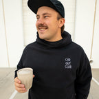 Man wearing a black hoodie with 'Car Guy Club' text, holding a coffee cup, standing in a parking lot.