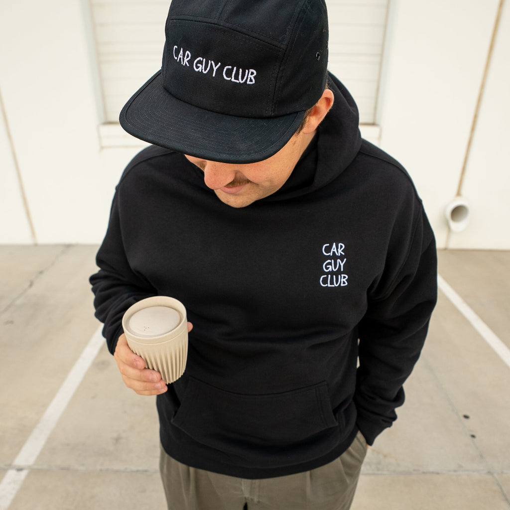 Person wearing a black 'Car Guy Club' hoodie and cap, holding a coffee cup.