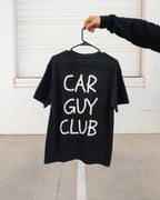 Black t-shirt with 'CAR GUY CLUB' text on a hanger against a white wall.