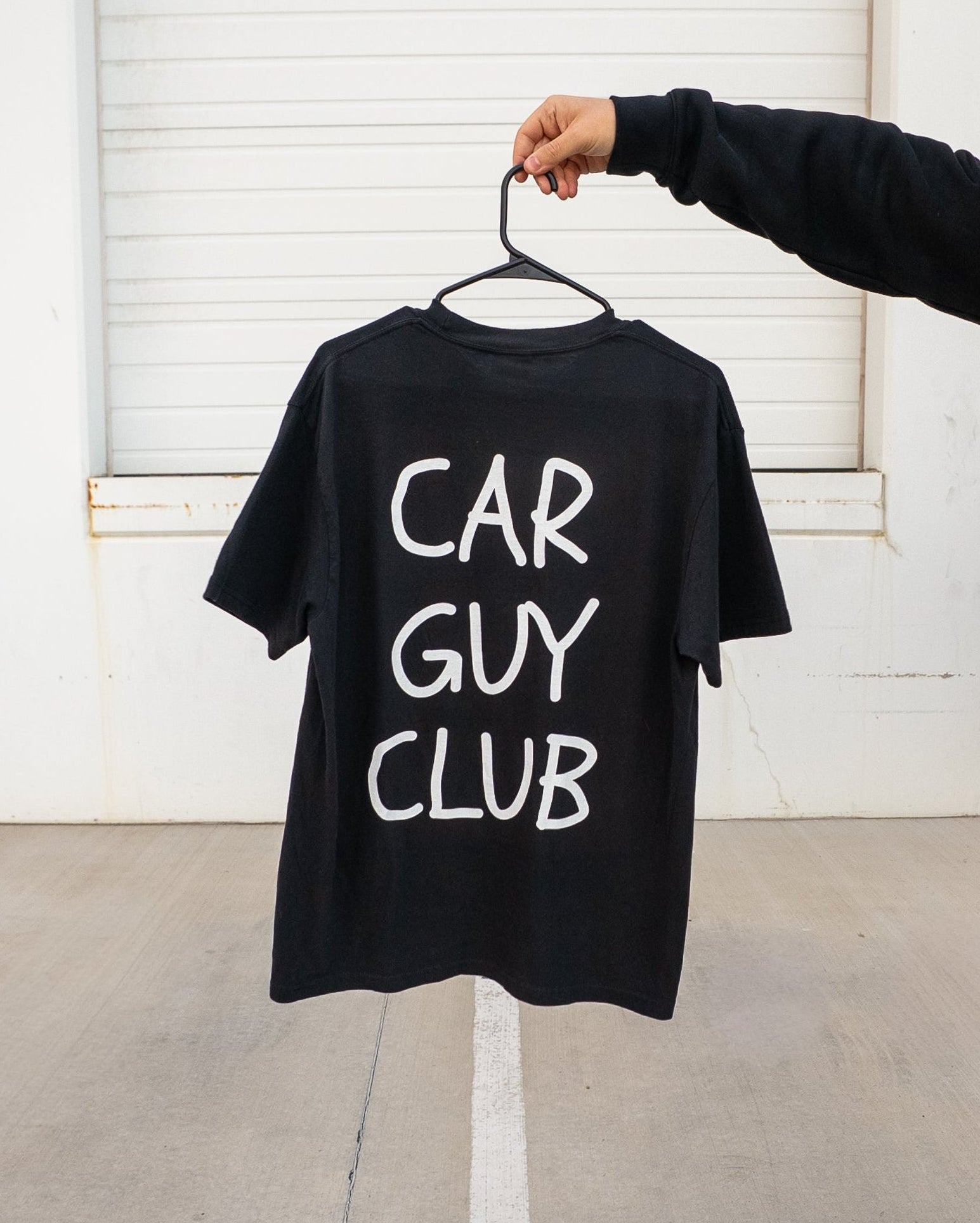 Black t-shirt with 'CAR GUY CLUB' text on a hanger against a white wall.