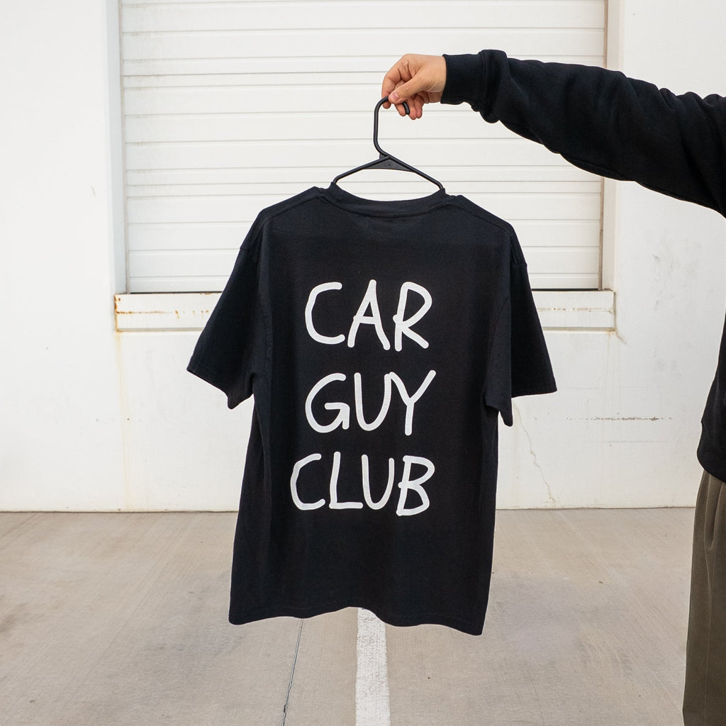 Person holding a black t-shirt with 'CAR GUY CLUB' text on a hanger against a white wall.