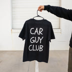 Person holding a black t-shirt with 'CAR GUY CLUB' text on a hanger against a white wall.