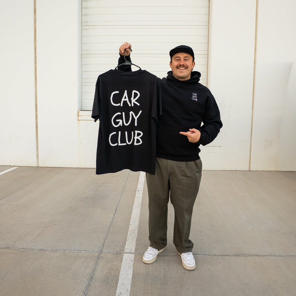 Person holding a 'Car Guy Club' t-shirt in front of a white wall.