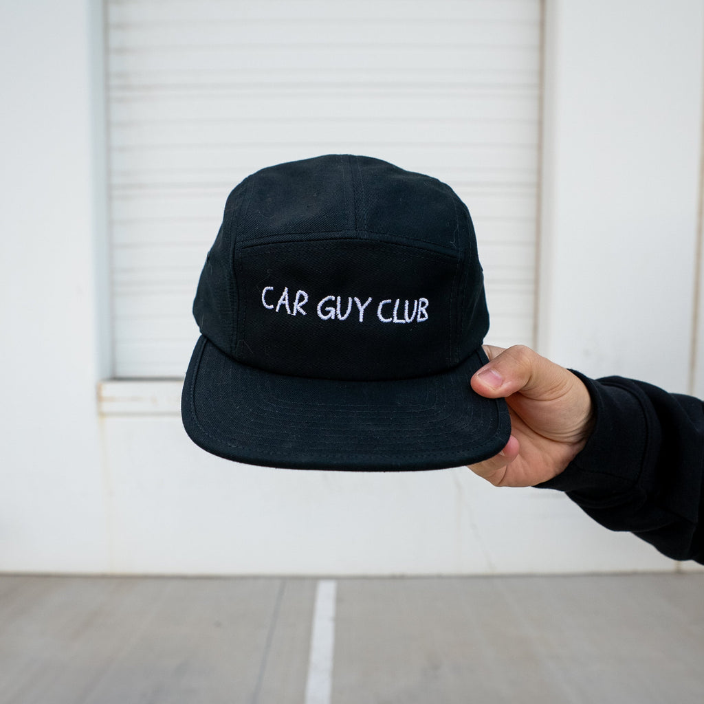 Black cap with 'CAR GUY CLUB' text held by a person against a white garage door.