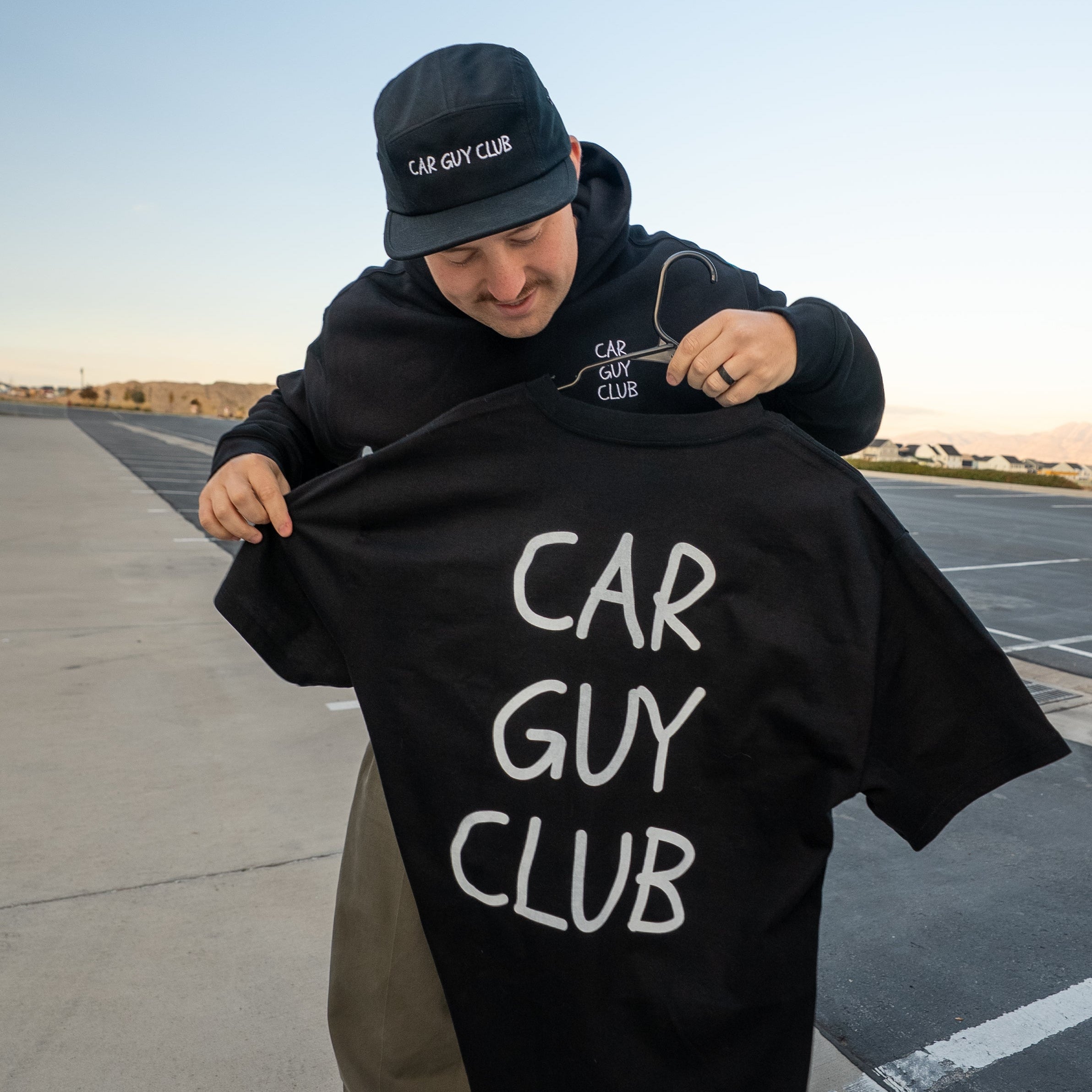 Person holding a black hoodie with 'CAR GUY CLUB' text on a paved road.