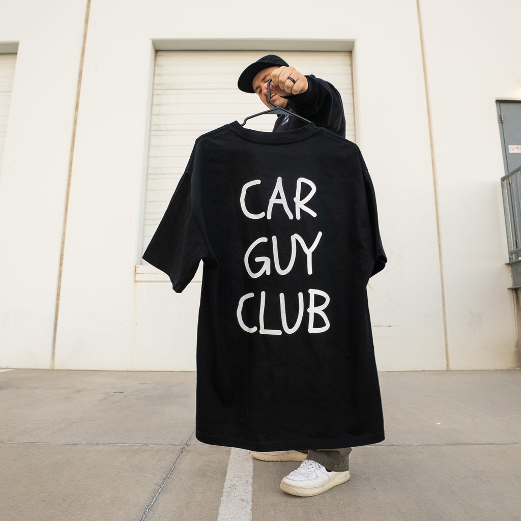 Person holding a black t-shirt with 'CAR GUY CLUB' text against a white wall.