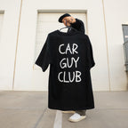 Person holding a black t-shirt with 'CAR GUY CLUB' text against a white wall.