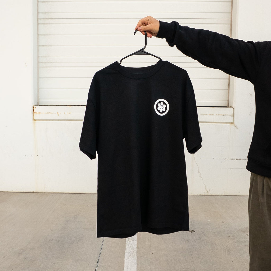 Black t-shirt with a white logo on a hanger against a white wall.