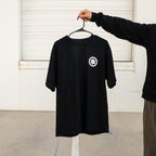 Black t-shirt with a white logo on a hanger against a white wall.