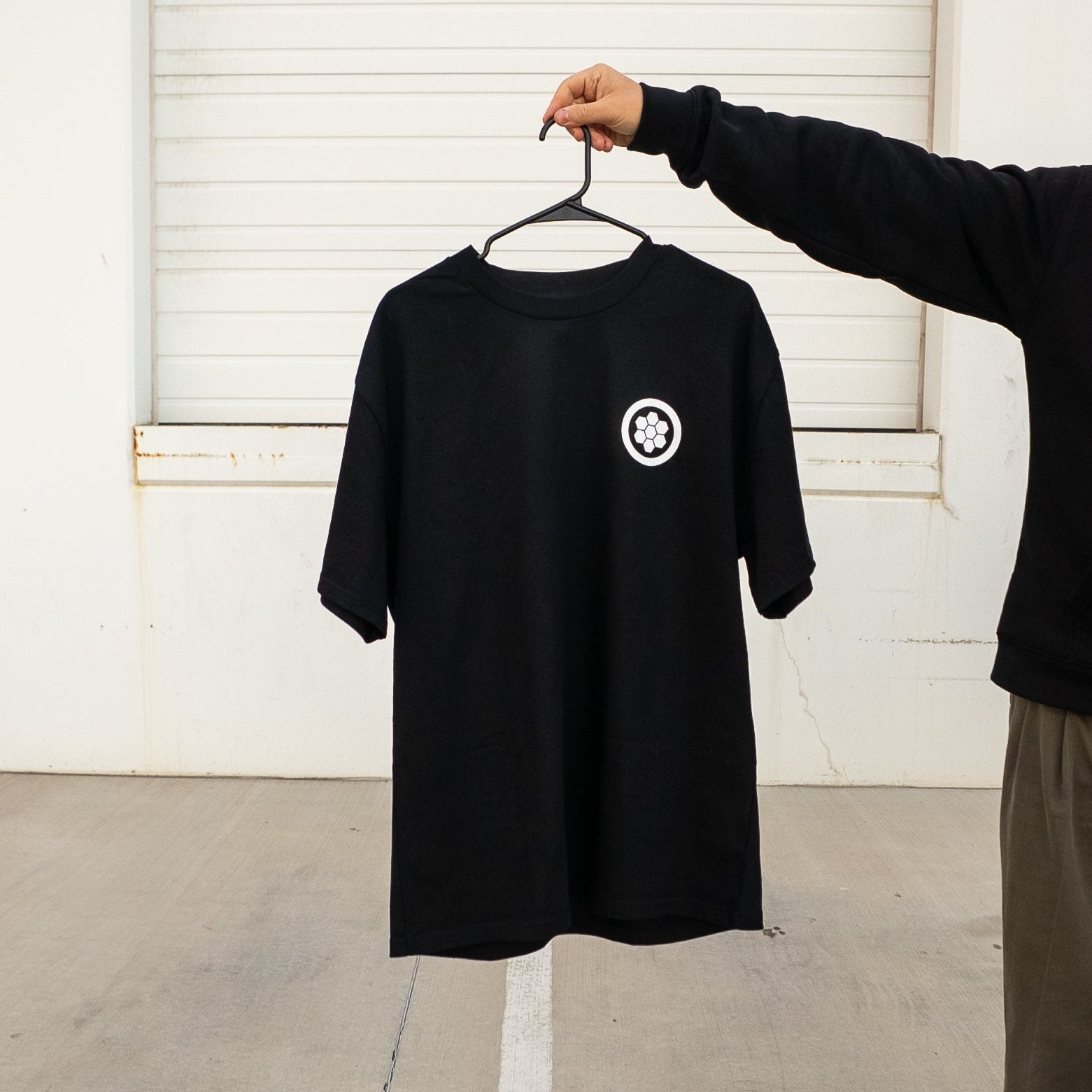 Black t-shirt with a white logo on a hanger against a white wall.