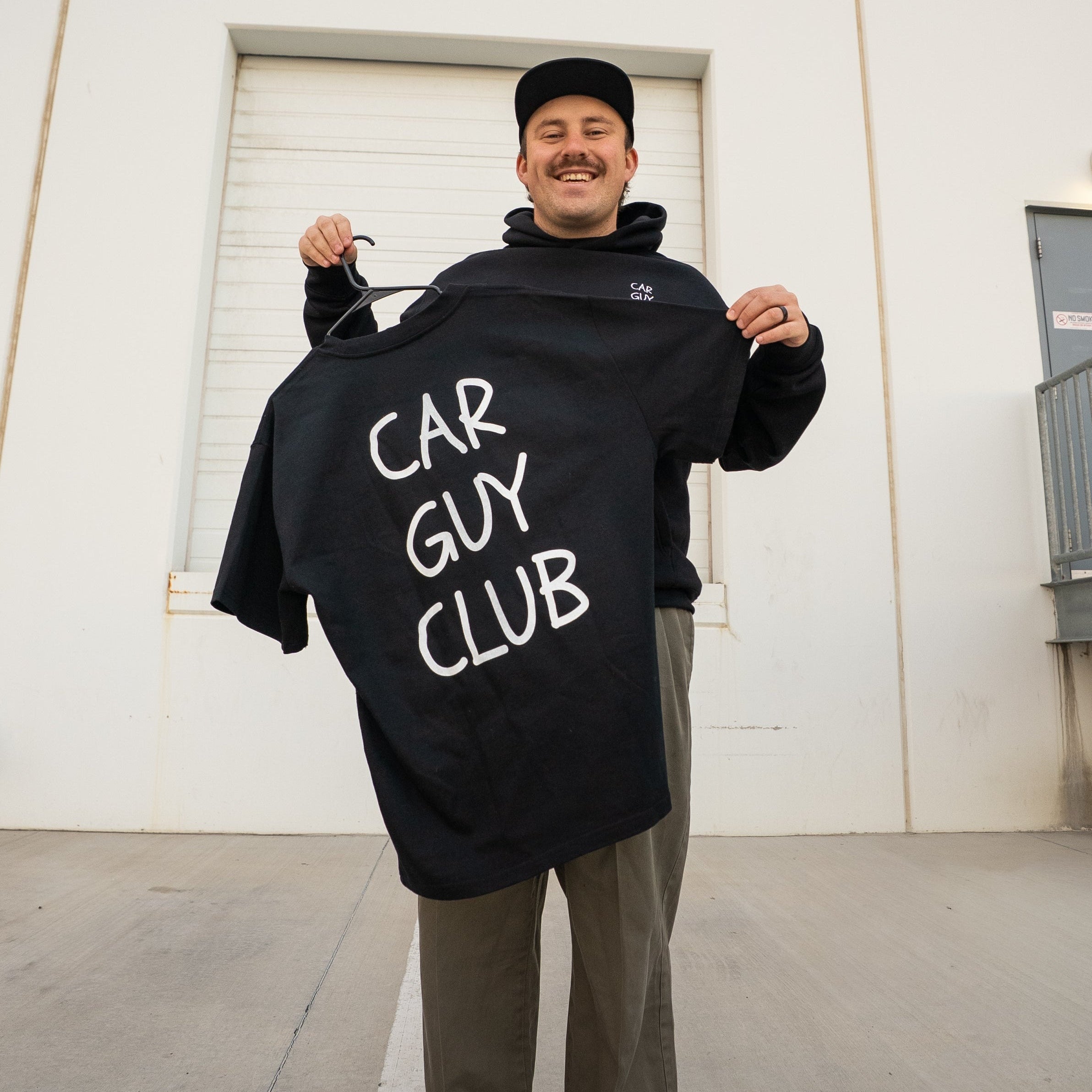 Person holding a black hoodie with 'CAR GUY CLUB' text against a white wall.