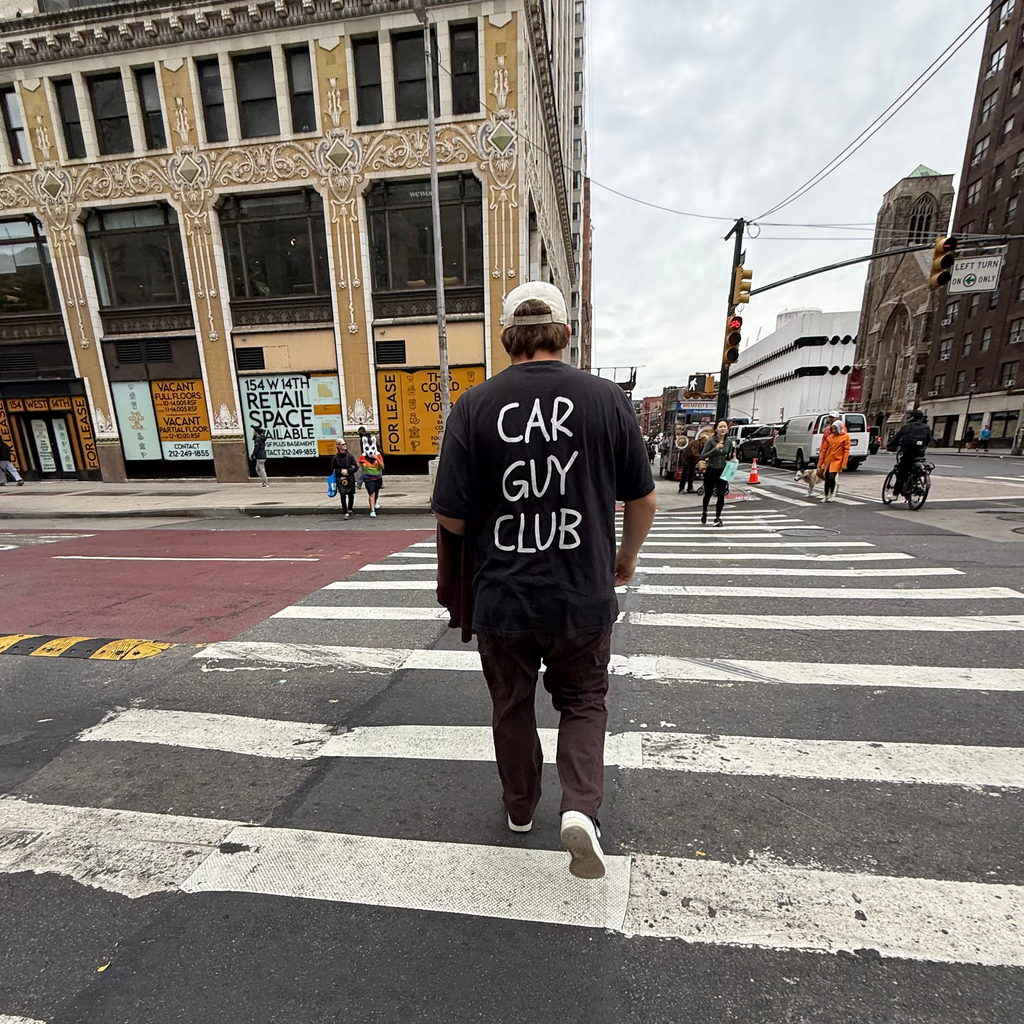 Person wearing a 'Car Guy Club' t-shirt crossing a street in an urban setting.